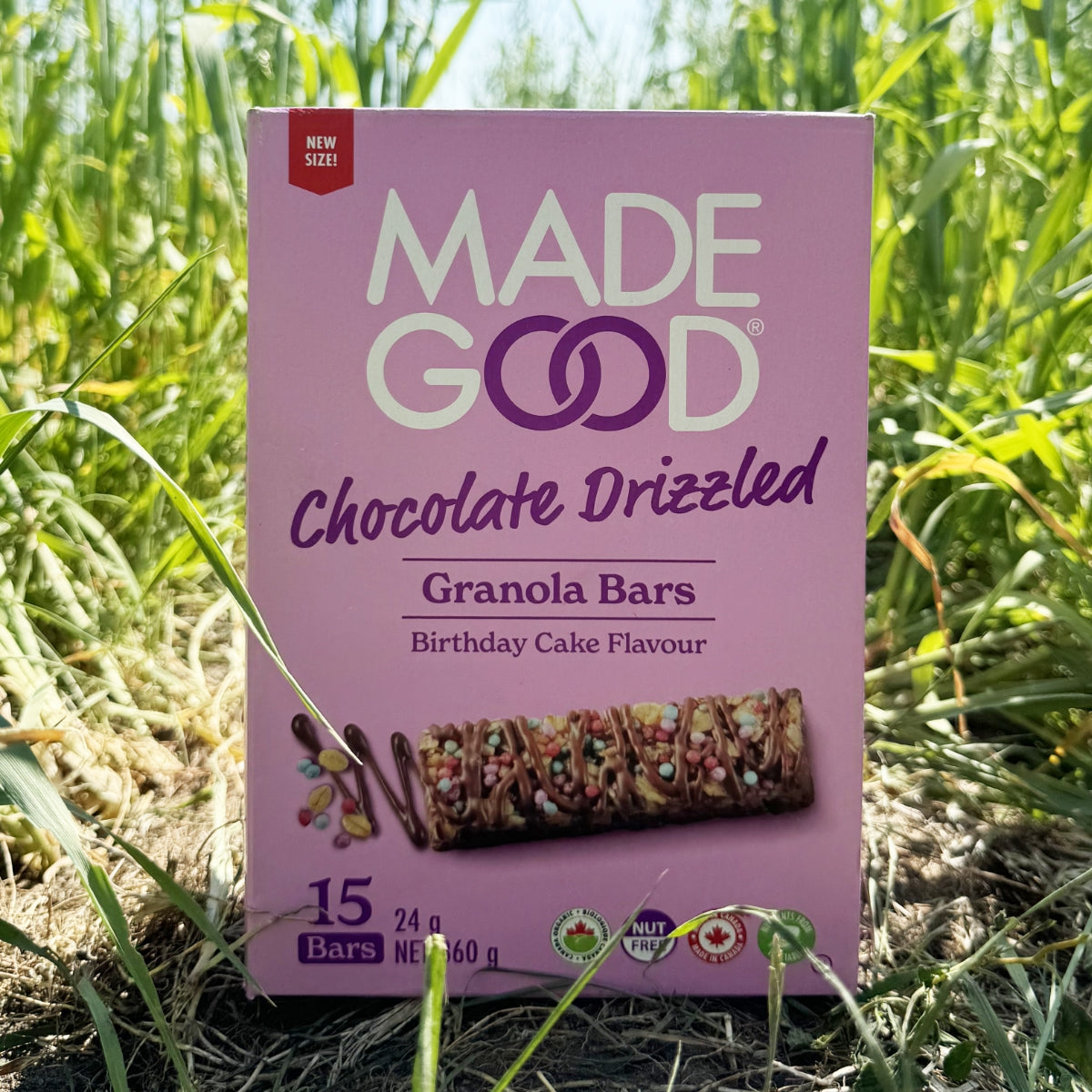 Box of Made Good Chocolate Drizzled Granola Bars, Birthday Cake flavor, in a grassy field Box of Made Good Chocolate Drizzled Granola Bars, Birthday Cake flavor, in a grassy field. The package is purple with bright, colorful accents.