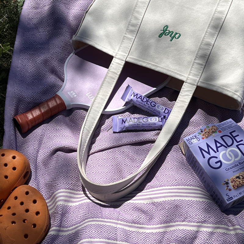 Cream tote bag, pickleball paddle, purple snack bars, and granola bar box on a lavender geometric-patterned blanket beside orange clogs. Sunny, relaxed picnic vibe.