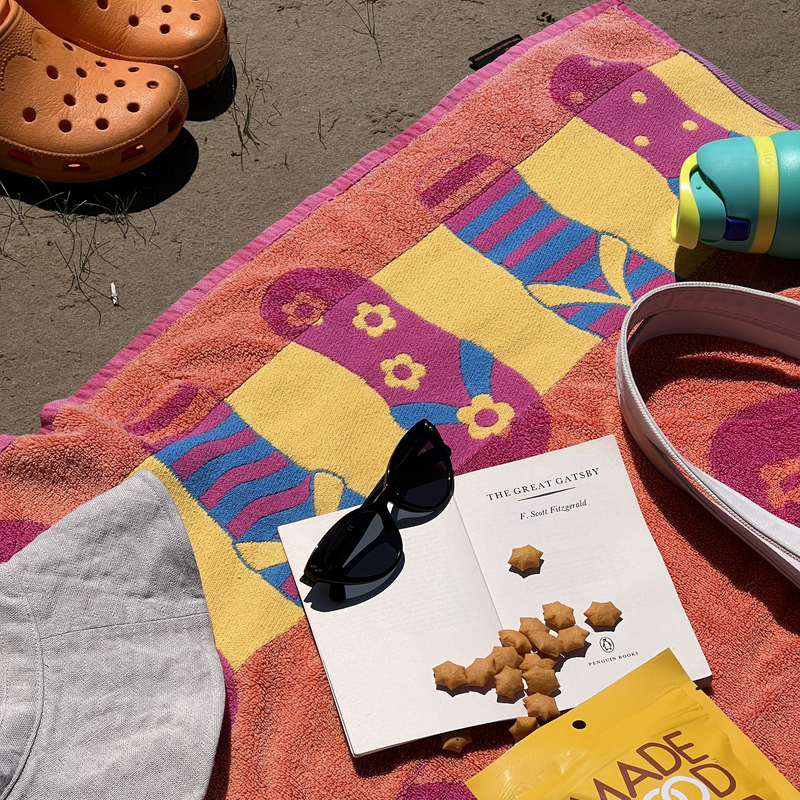 Beach scene with colorful towel, orange clogs, sunglasses on an open book, a water bottle, snacks, and a hat. Relaxed and sunny atmosphere.
