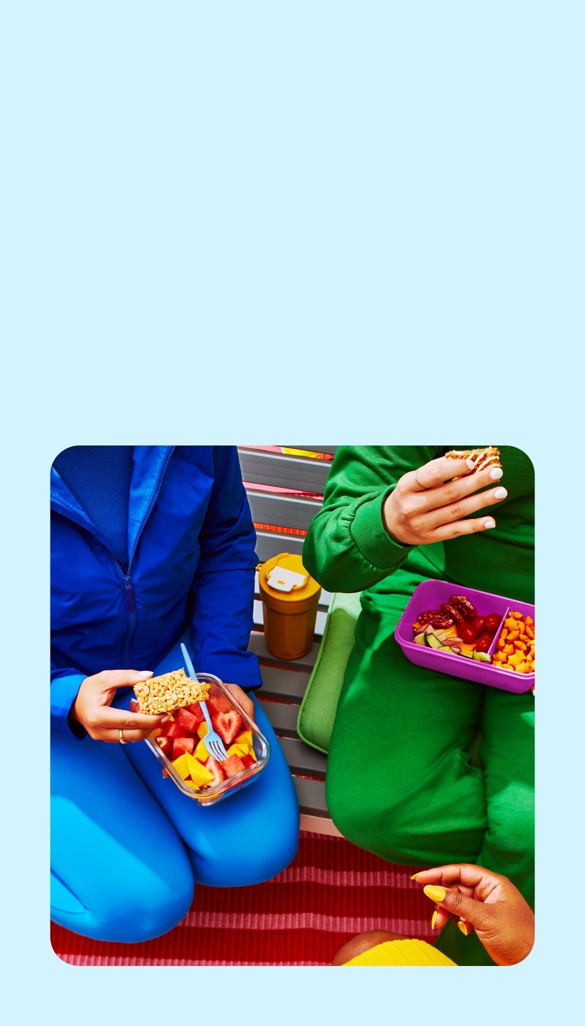 Two people sit side-by-side, one in blue with a fruit bowl and coffee cup, the other in green with a purple tray of strawberries and snacks. Casual and relaxed setting.